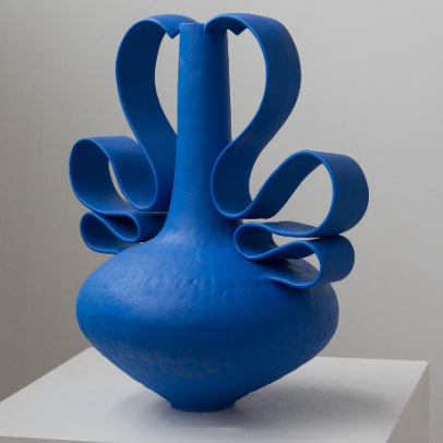 The Wave Vessel Cobalt by Latch Key Ceramics is a cobalt blue ceramic piece featuring a round base and wavy, ribbon-like extensions looping from its neck, elegantly displayed on a white pedestal.