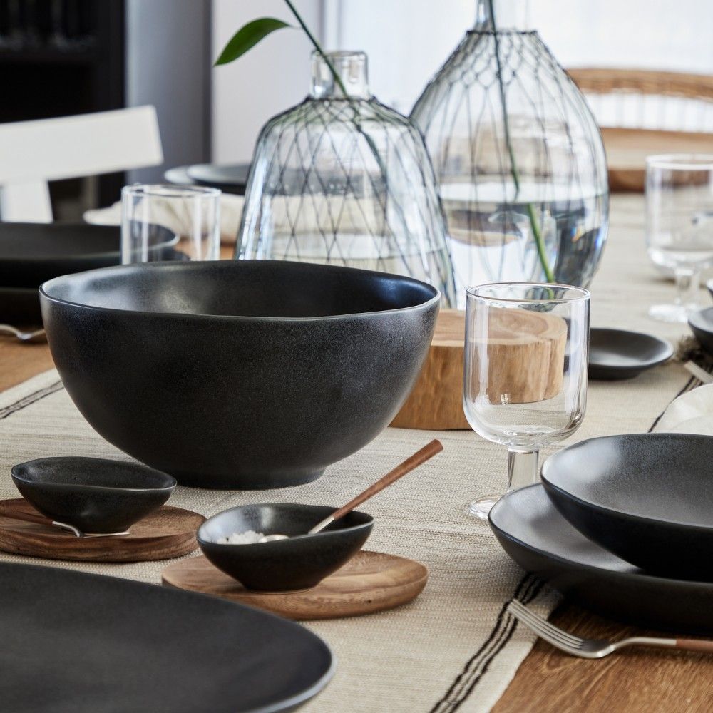 A dining table set with modern flair, featuring Casafina/Costa Nova's Livia Dinnerware Matte Black stoneware bowls and plates made in Portugal on wooden trays, clear glassware, glass vases, and a striped table runner.