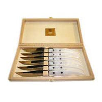 The Claude Dozorme Berlingot Steak Knives Set of 6 comes in a wooden box with a cream interior. Six steak knives, each with a shiny blade and white handle featuring two black dots, are neatly arranged inside.
