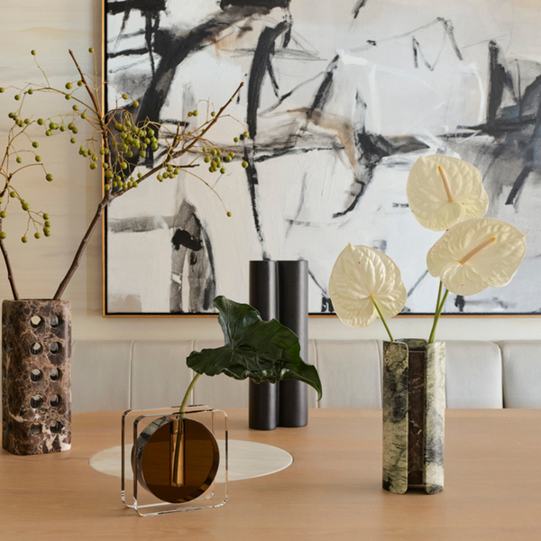 A modern table displays the Greg Natale Caldera Crystal Vase - Clear & Umber, filled with green branches, leafy stems, and white anthurium flowers. Abstract black and white art hangs on the wall behind this sculptural arrangement.