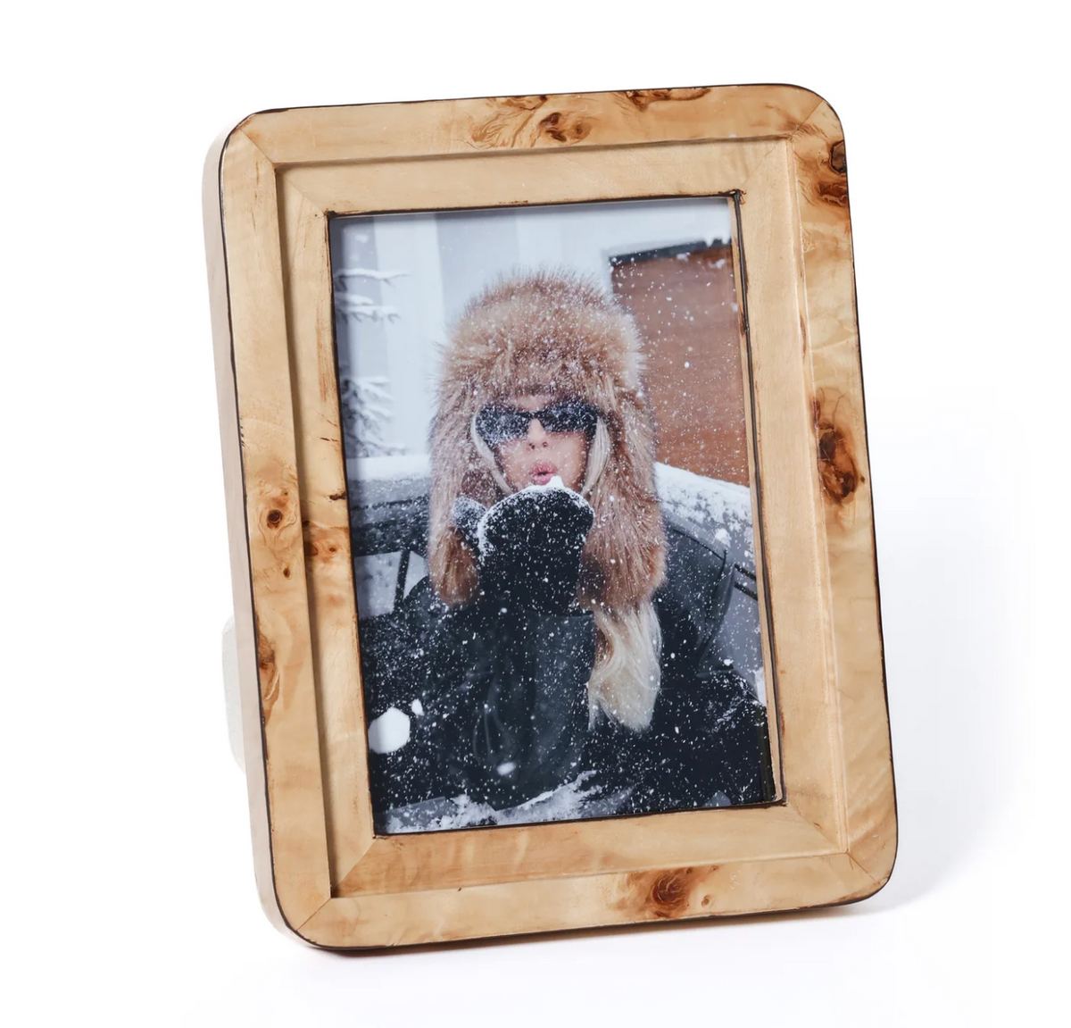 The Valari Burl Veneer Frame by Zodax showcases a photo of a person in winter attire—furry hat and sunglasses—blowing snow from their hands, with snowflakes visible in the air.