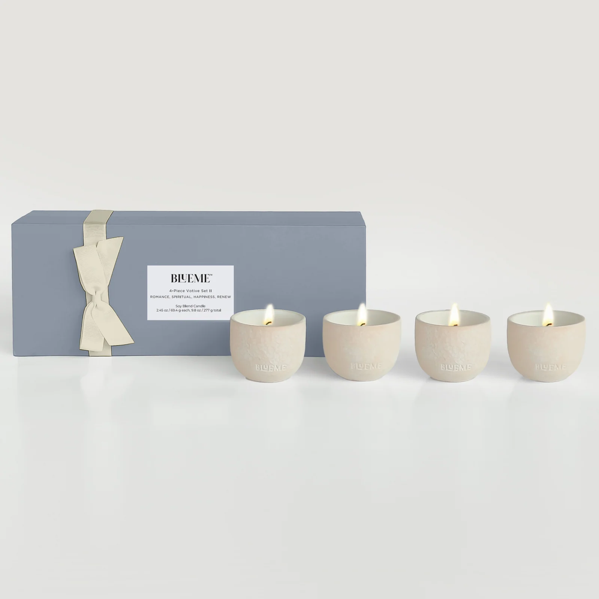 The Blueme Travel Votive 4 Piece Set—Romance, Spiritual, Happiness, Renew—features four lit cream soy candles in handcrafted ceramic holders beside a gray gift box with a cream ribbon on a white surface.