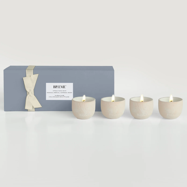 The Blueme Travel Votive 4 Piece Set—Romance, Spiritual, Happiness, Renew—features four lit cream soy candles in handcrafted ceramic holders beside a gray gift box with a cream ribbon on a white surface.