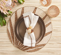 A brown plate is topped with a Kim Seybert Filament Napkin in white and brown, elegantly held by a beaded napkin ring and set on a woven placemat with brown rays. Pink flowers and two glasses are positioned at the top left and right, completing this sophisticated tablescape featuring the Filament Napkin Set of 4 by Kim Seybert.