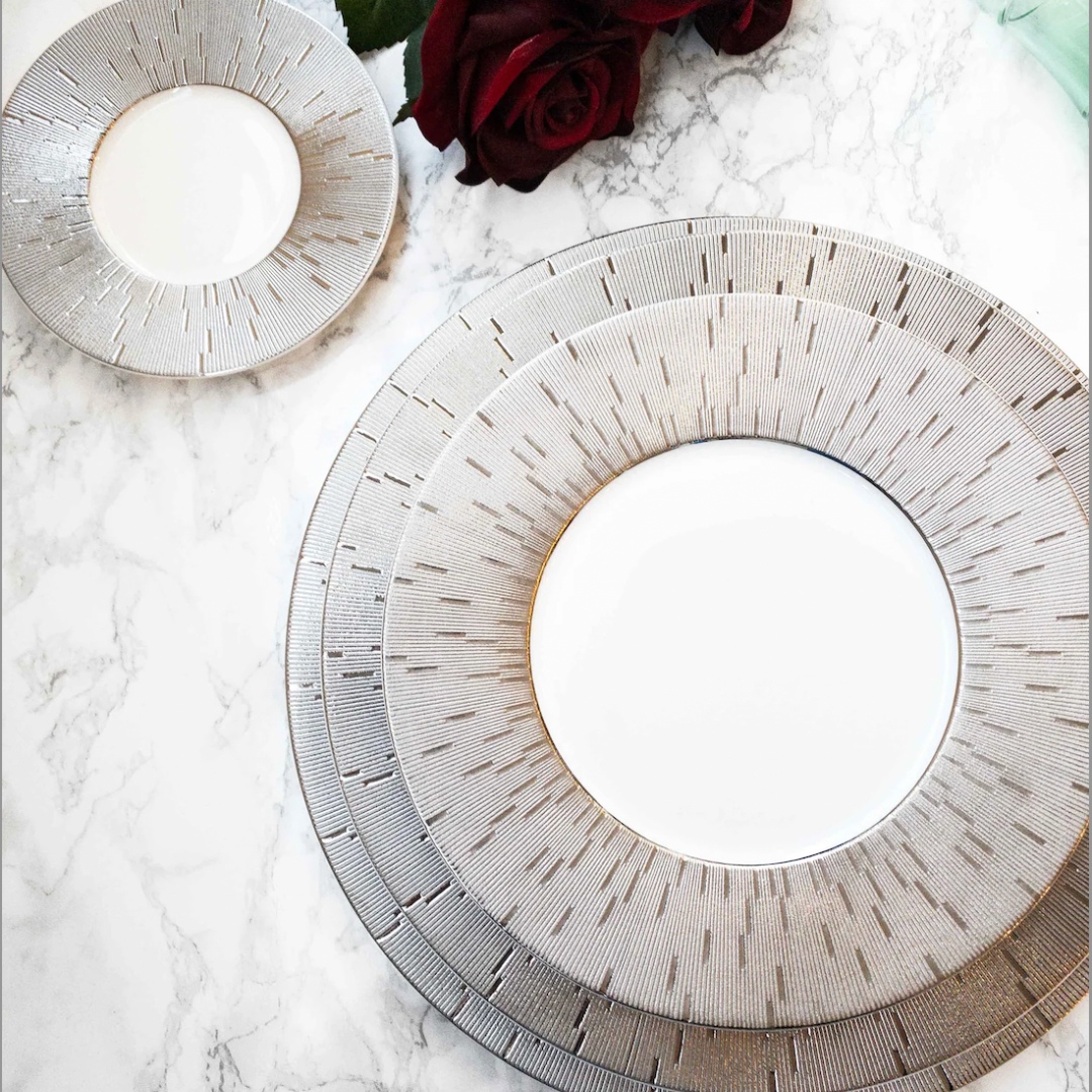 Two Infini Prestige Platinum Dinnerware plates by Haviland, crafted from elegant white Limoges porcelain with intricate platinum detailing, rest on marble. A deep red rose and green leaves add a sophisticated touch to the dining scene.