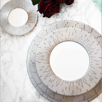 Two Infini Prestige Platinum Dinnerware plates by Haviland, crafted from elegant white Limoges porcelain with intricate platinum detailing, rest on marble. A deep red rose and green leaves add a sophisticated touch to the dining scene.