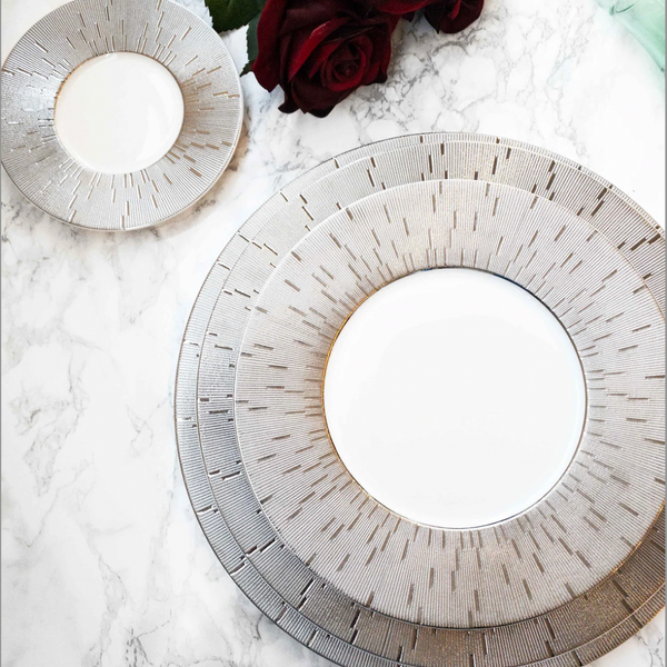 Two Infini Prestige Platinum Dinnerware plates by Haviland, crafted from elegant white Limoges porcelain with intricate platinum detailing, rest on marble. A deep red rose and green leaves add a sophisticated touch to the dining scene.