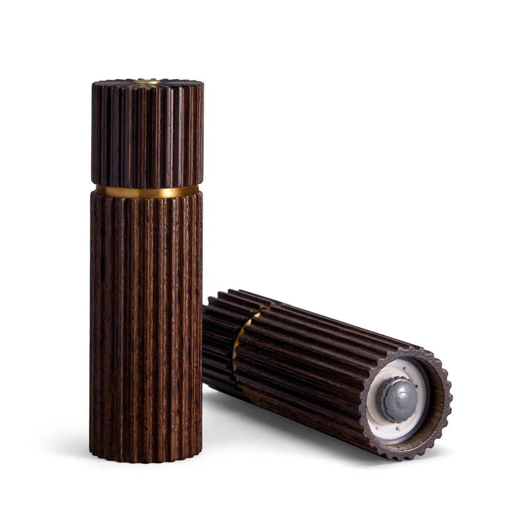 The L’Objet Ionic Salt & Pepper Mill Smoked Oak Set of 2 features cylindrical mills with ribbed, smoked oak exteriors and brass accents; one is upright, the other is horizontal, showing a clear opening on one end.