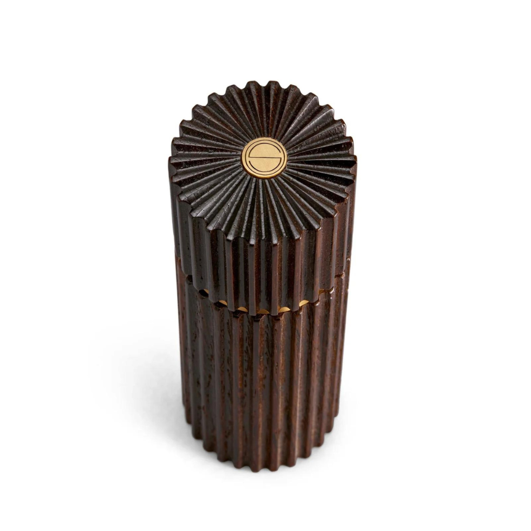 The L'Objet Ionic Salt & Pepper Mill Smoked Oak Set of 2 features dark, ridged cylindrical mills with flower-like tops and gold emblems. Made from European oak, they include a durable metal grinding mechanism and are shown on a white background.