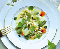 The Bernardaud Palmyre Dinnerware showcases neatly arranged leeks topped with diced vegetables, herbs, and edible flower petals. A gold fork and green leaf complete the refined, elegant table presentation.