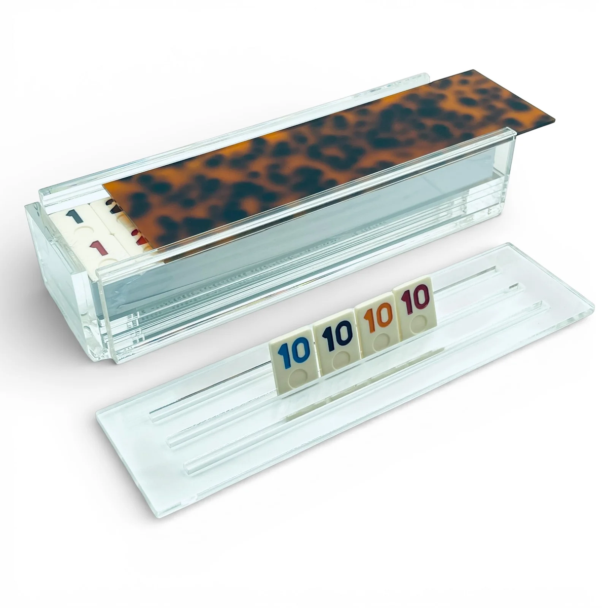 The Luxe Dominoes Rumi Game - Tortoise features numbered game tiles stored in a clear acrylic box with a tortoiseshell lid. In front, four tiles—each displaying the number 10 in different colors—are arranged on a transparent tile holder.