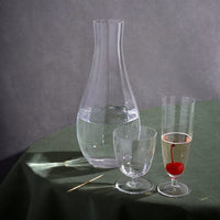 The Iris Drinkware Collection by L'Objet, featuring a mouth-blown glass carafe, goblet, and champagne flute with a cherry garnish, is elegantly displayed on a green tablecloth against a gray backdrop.