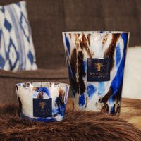 Two Baobab Halona Candle Blue World candles, presented in marbled blue, brown, and white hand-blown glass holders, rest on a furry brown surface with a patterned cushion and sofa in the background.
