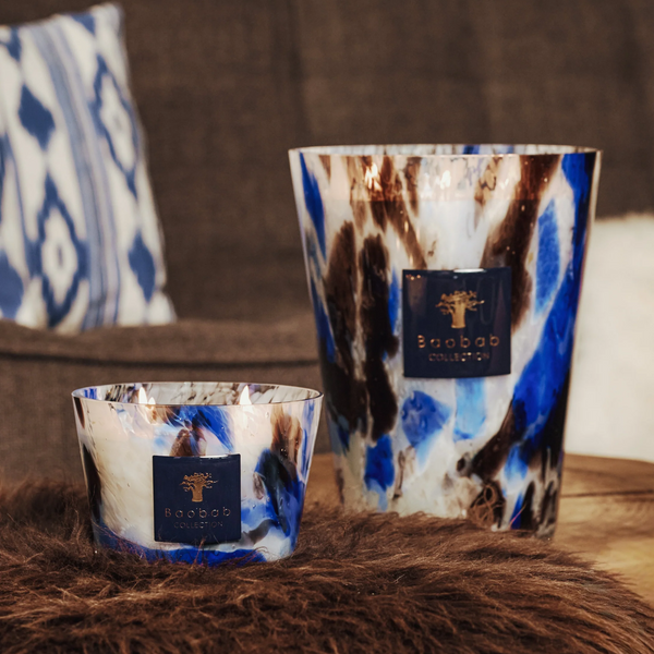 Two Baobab Halona Candle Blue World candles, presented in marbled blue, brown, and white hand-blown glass holders, rest on a furry brown surface with a patterned cushion and sofa in the background.