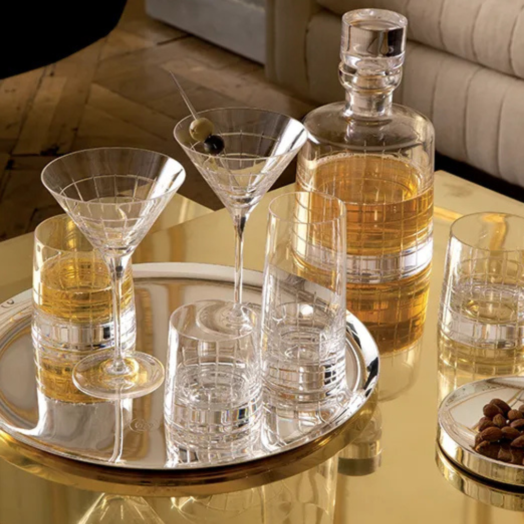 A reflective gold table displays the Christofle Graphik Barware Set of 2 by Christofle, alongside crystal glassware, a decanter with amber liquor, martini glasses (one with an olive), extra glasses, a nut bowl, and elegant silver barware.
