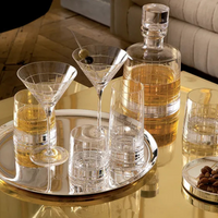 A reflective gold table displays the Christofle Graphik Barware Set of 2 by Christofle, alongside crystal glassware, a decanter with amber liquor, martini glasses (one with an olive), extra glasses, a nut bowl, and elegant silver barware.