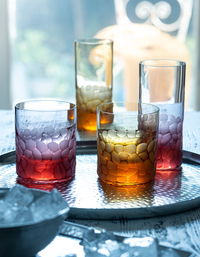 Four Moser Pebbles DOF 370 ml hand-blown glass tumblers with red-to-gold gradients sit on a silver tray as sunlight streams in. A bowl of ice is in the foreground, ideal for enjoying whisky on a white wooden surface.