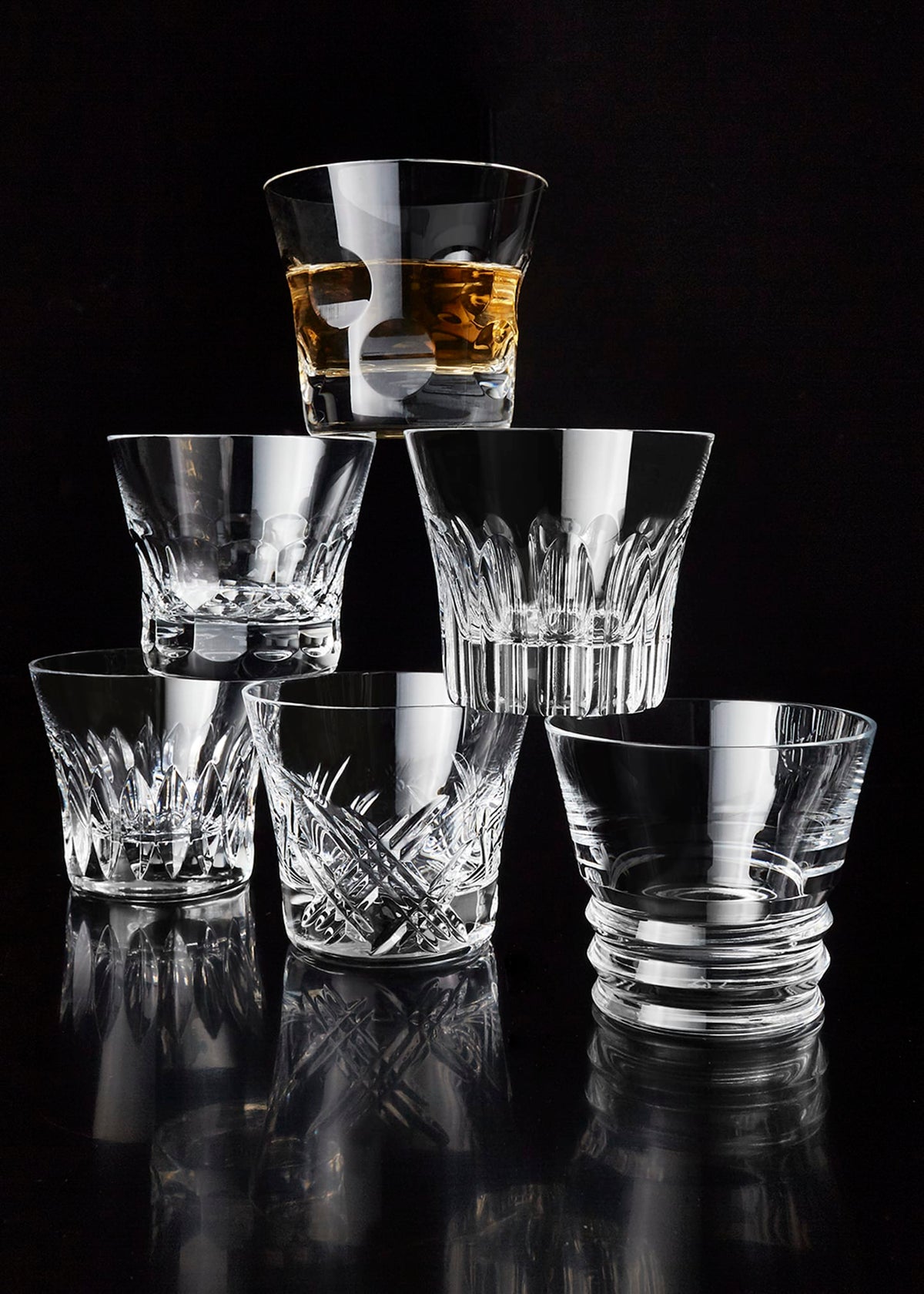 Six Everyday Tumblers by Baccarat are stacked in a pyramid against a black background. The top glass holds whiskey, while the rest are empty, showcasing their elegance—ideal for presenting as a luxury gift set.