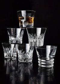 Six Everyday Tumblers by Baccarat are stacked in a pyramid against a black background. The top glass holds whiskey, while the rest are empty, showcasing their elegance—ideal for presenting as a luxury gift set.