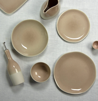 A flat lay of beige Maguelone Dinnerware Tamaris by Jars - Rosenthal, featuring plates, a serving bowl, bowls, a pitcher, a small cup, and an olive oil bottle on white linen with subtle crackled glazes.