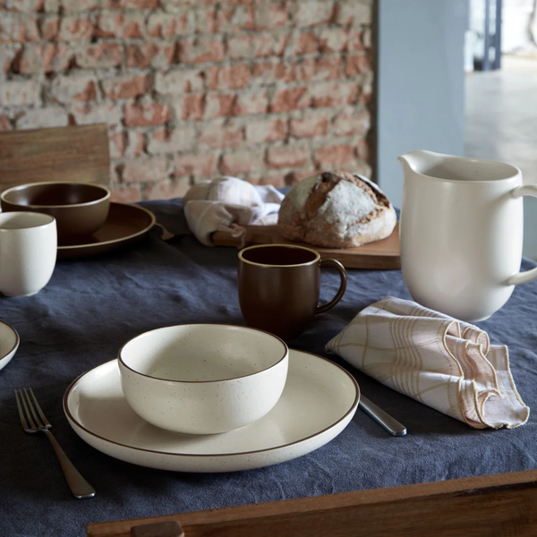 A rustic table setting featuring Casafina/Costa Nova Pacifica Dinnerware Monterosa Beige, with a pitcher, cups, a loaf of bread, cutlery, and checkered napkins on a blue tablecloth against a brick wall.