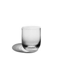 The Optic Glassware Clear Set of 2 by Richard Brendon features ribbed glass tumblers with a diagonal swirl pattern, shown on a white surface casting a patterned shadow to the left.
