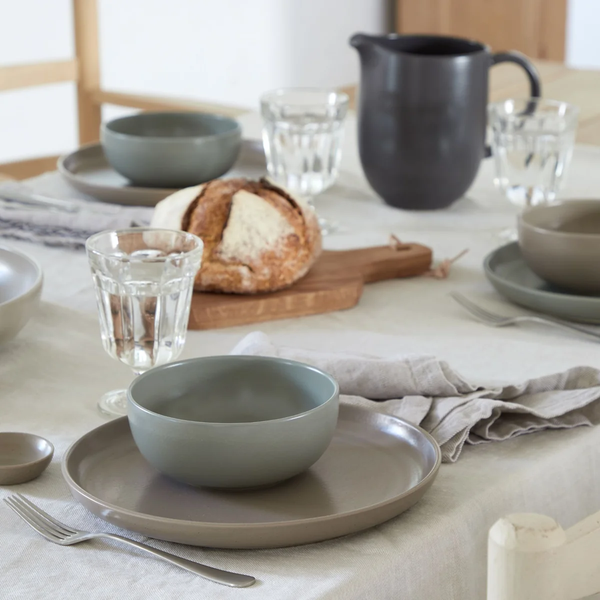 A table set for a meal with Costa Nova’s Pacifica Dinnerware in Artichoke, featuring gray plates and bowls, glasses of water, a loaf of bread on a wooden board, napkins, and a dark pitcher atop a light tablecloth, exudes the charm of elegant dinnerware.