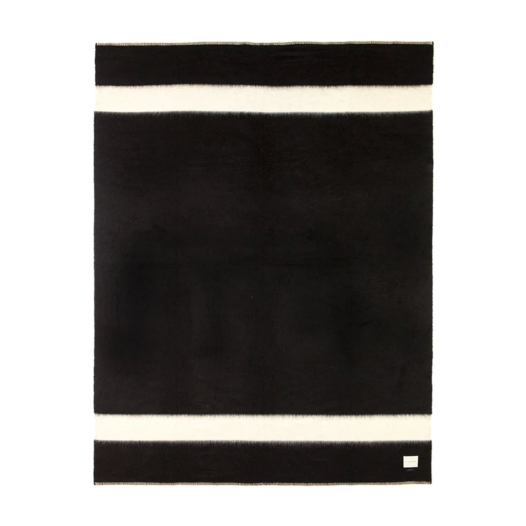 The Siempre Recycled Alpaca Throw by Blacksaw features a rectangular black design with two bold white stripes at the top and bottom edges, plus a small white label in one corner. This heavyweight blanket is perfect for cozy fireside evenings.