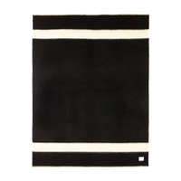 The Siempre Recycled Alpaca Throw by Blacksaw features a rectangular black design with two bold white stripes at the top and bottom edges, plus a small white label in one corner. This heavyweight blanket is perfect for cozy fireside evenings.