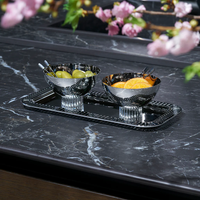 The Striations Silver Nut Bowl by Anna New York, filled with green grapes, sits on a reflective tray atop a dark marble counter. Another silver bowl holds orange slices, while pink blossoms blur softly in the foreground.