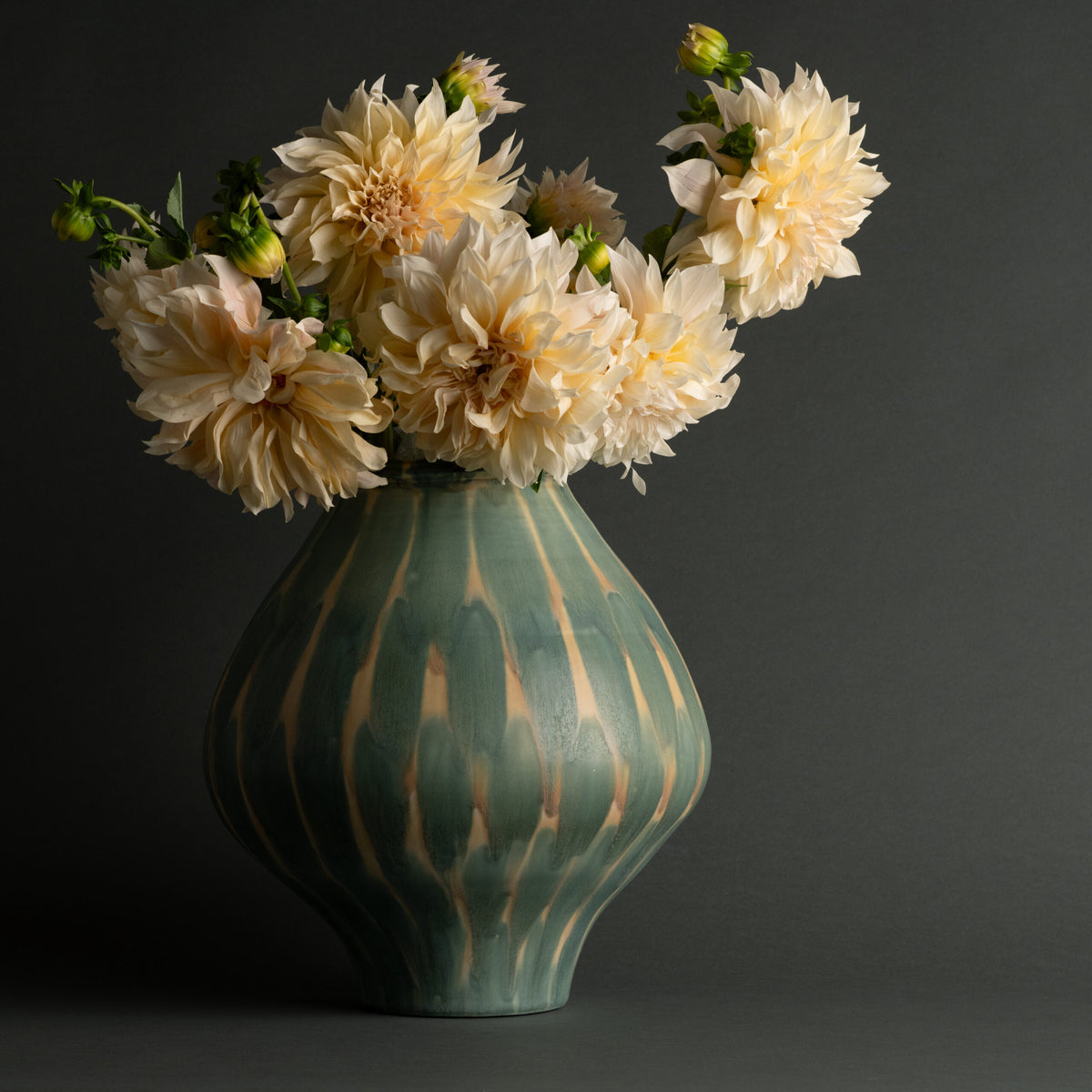 The Jace Vase by FlowDecor Inc., a decorative green and beige striped ceramic piece, is filled with large, pale peach dahlias and set against a dark gray background.