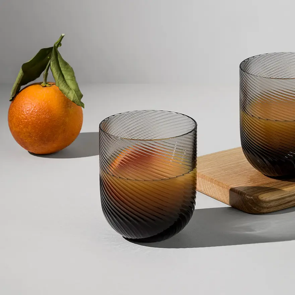 A glass from the Optic Glassware Smoke Set of 2 by Richard Brendon, filled with orange juice, sits on a gray surface near a cutting board with an orange in the background, showcasing its stylish contemporary design.