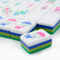 A close-up of Oh My Mahjong’s Jasmine Mahjong Tiles Set shows a vibrant acrylic tile with a blue monkey, pink umbrella, and “JOKER.” The tiles feature green, blue, and yellow layered sides.