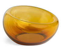 The Axiom Glass Caldera Vessel Medium 9" is a round, hand-blown glass piece with thick, translucent amber walls and a slightly tilted rim, displayed on a white background.
