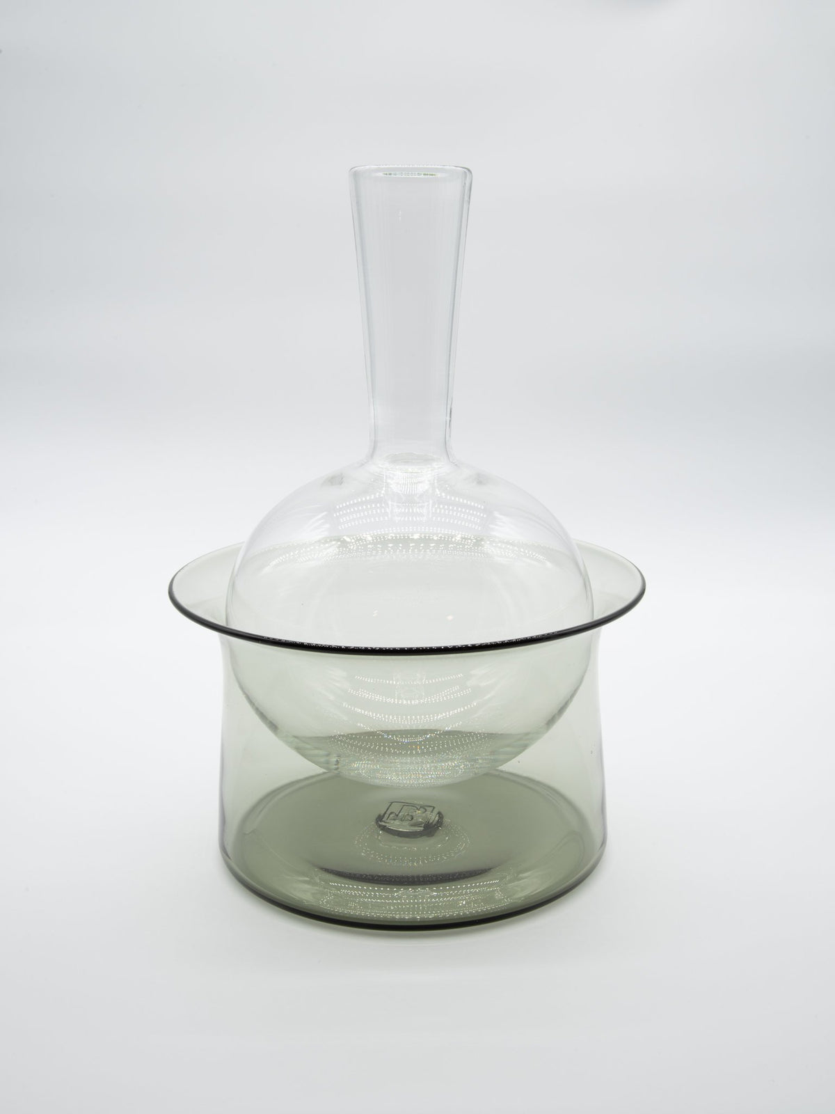 The Equilbrium Decanter - Smoke by Tyler Barry Design, featuring a long neck and round base, sits inside a wide, shallow cylindrical glass bowl against a plain white background.