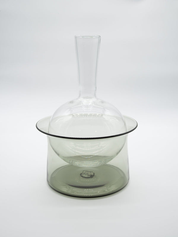 The Equilbrium Decanter - Smoke by Tyler Barry Design, featuring a long neck and round base, sits inside a wide, shallow cylindrical glass bowl against a plain white background.