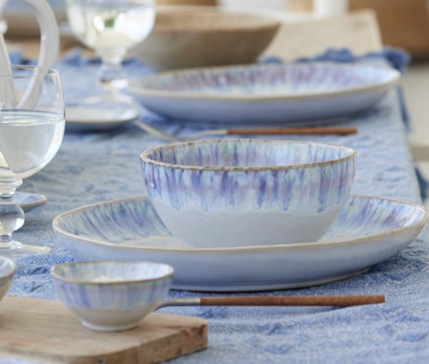 A close-up of Costa Nova's Brisa Blue Dinnerware, crafted in Portugal, featuring blue and white patterns on a table with water glasses, a blue tablecloth, and wooden chopsticks.