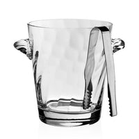 The William Yeoward Dakota Ice Bucket features clear glass and textured side handles, paired with metal ice tongs on the rim. Perfect for your wet bar or as a housewarming gift. The empty bucket is displayed against a white background.