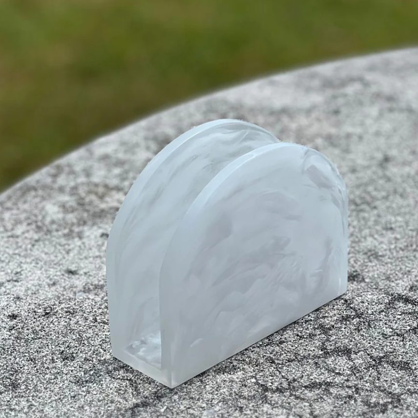 The BT-Shalom Dawn Resin Swirl Napkin Holder, featuring a white marble pattern, stands upright on a speckled gray stone surface and complements modern kitchen décor. Blurred green grass can be seen in the background.