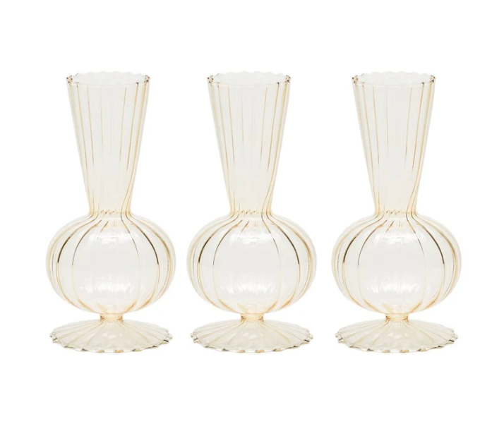 Four identical champagne-colored glass bud vases from Kim Seybert's Tess Bud Vase Set, featuring vertical ribbed patterns, rounded bases, and fluted edges, are arranged in a row against a white background.