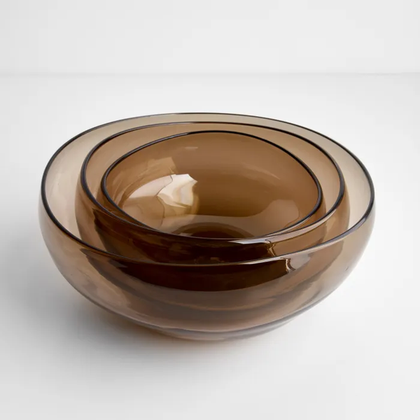 The Gary Bodker Nesting Glass Bowl Collection Wheat features three handblown translucent brown bowls in varied sizes, beautifully stacked on a white surface—a stylish set ideal for serving or use as salad bowls.