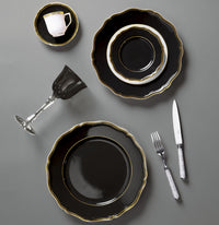 A formal table setting with Raynaud's Mazurka Dinnerware Or Black—black and gold-edged plates, a matching cup and saucer, plus a clear wine glass—exudes luxury and showcases exquisite French craftsmanship on a refined grey surface.