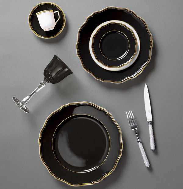 A formal table setting with Raynaud's Mazurka Dinnerware Or Black—black and gold-edged plates, a matching cup and saucer, plus a clear wine glass—exudes luxury and showcases exquisite French craftsmanship on a refined grey surface.