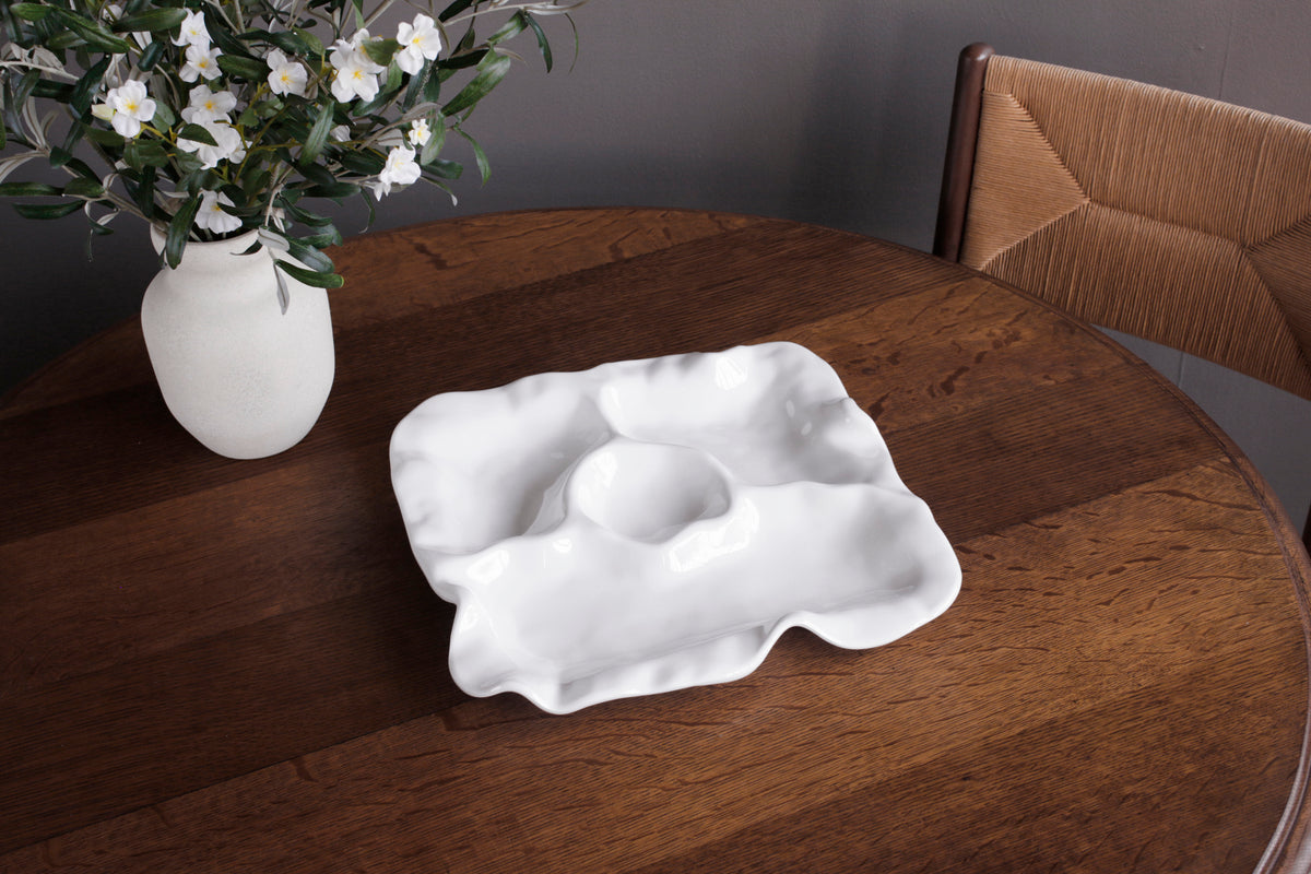 The Beatriz Ball Vida Havana Multi Chip & Dip, a white divided serving dish with wavy edges, sits on a round wooden table by a vase of white flowers and a woven-back chair—perfect for outdoor entertaining.
