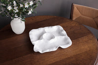 The Beatriz Ball Vida Havana Multi Chip & Dip, a white divided serving dish with wavy edges, sits on a round wooden table by a vase of white flowers and a woven-back chair—perfect for outdoor entertaining.