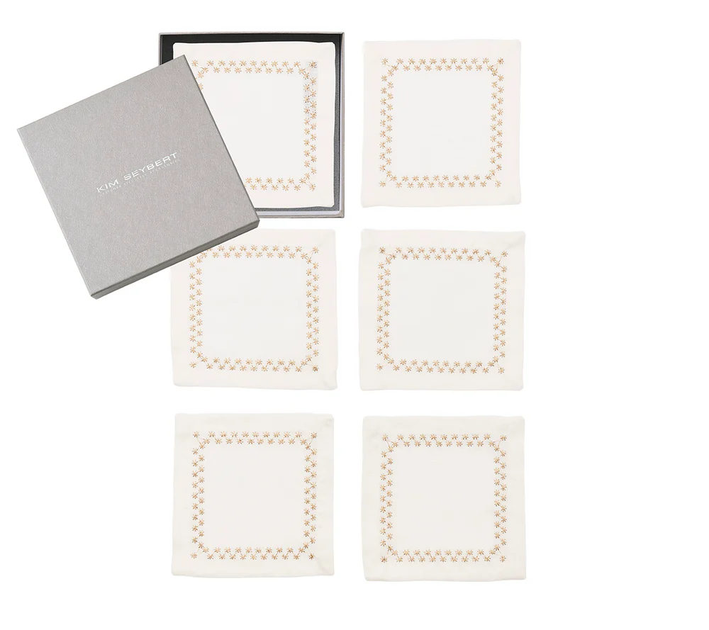 Six white Starry Night Cocktail Napkins by Kim Seybert, each with a delicate brown border, are arranged in two rows of three; an open gray Kim Seybert box is displayed above the top left napkin.