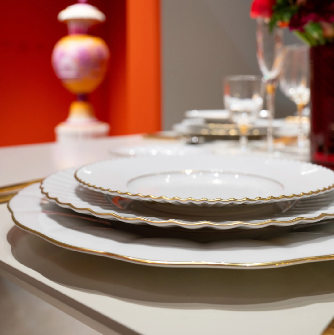 A close-up of a formal dining table set with Vista Alegre’s 200 Anos Dinnerware—stacked white plates with hand-painted gold trim—paired with glasses and cutlery. An orange wall and decorative vase appear in the background.