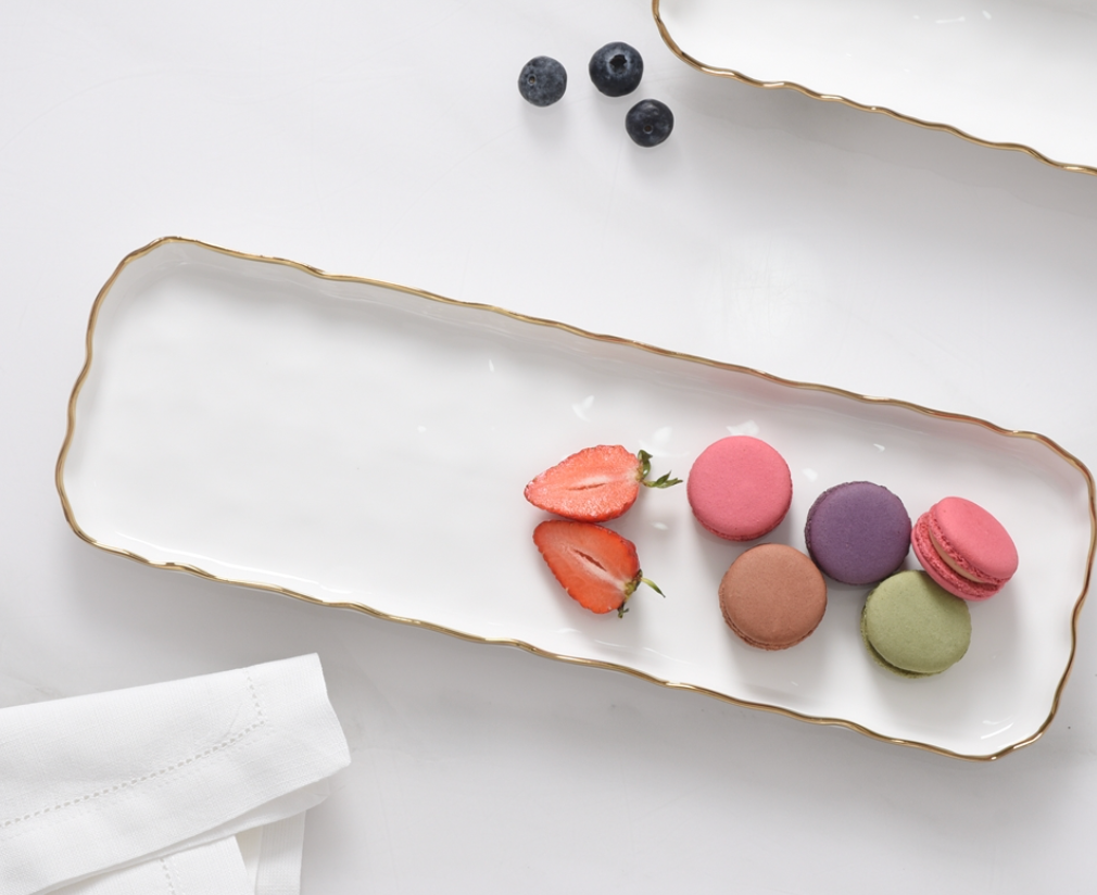 The PAMPA BAY Portofino Rectangle Tray with gold trim holds four colorful macarons and two strawberry halves. Three blueberries rest nearby on the table next to a white napkin, offering an elegant oven-to-table presentation.