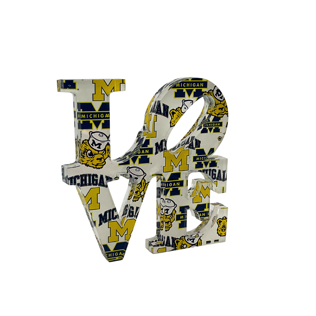 The Eye Candy College Love Sculpture is a clear acrylic piece featuring University of Michigan logos, mascot, and school colors—an ideal graduation gift for Wolverines fans or alumni.