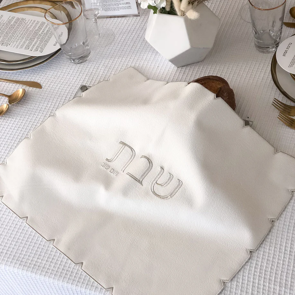 The BT-Shalom White Embroidered Leatherette Challah Cover- Silver is draped over challah bread on a white tablecloth, accented by gold utensils, plates, glasses, and a white vase for a stylish Shabbat meal setting.